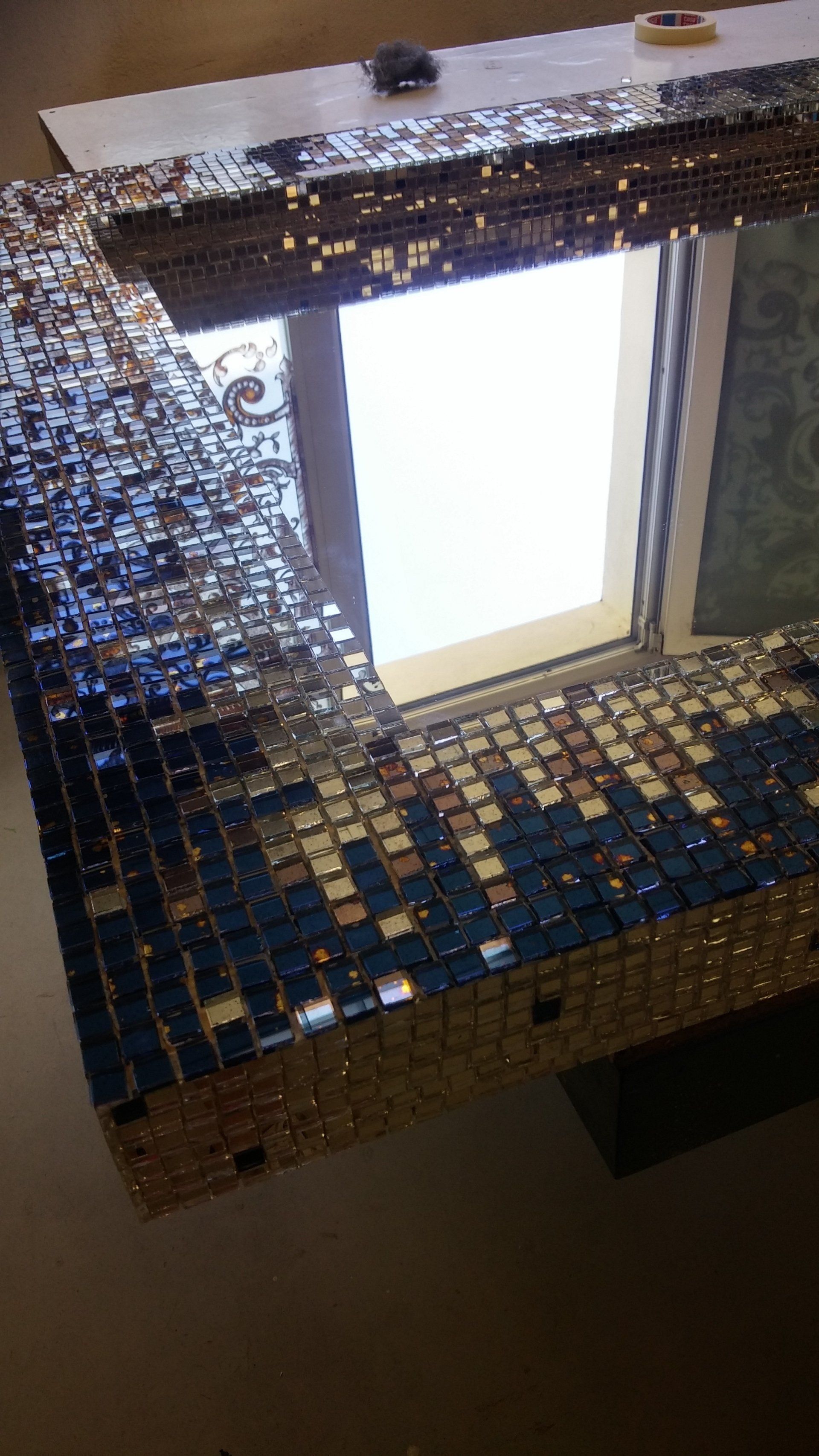 Cornice dello specchio con piastrelle a mosaico, blu, oro e argento; il riflesso mostra la finestra.