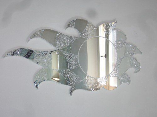 Specchio decorativo a forma di sole con centro circolare e raggi ondulati rivestiti di mosaico su una parete bianca.