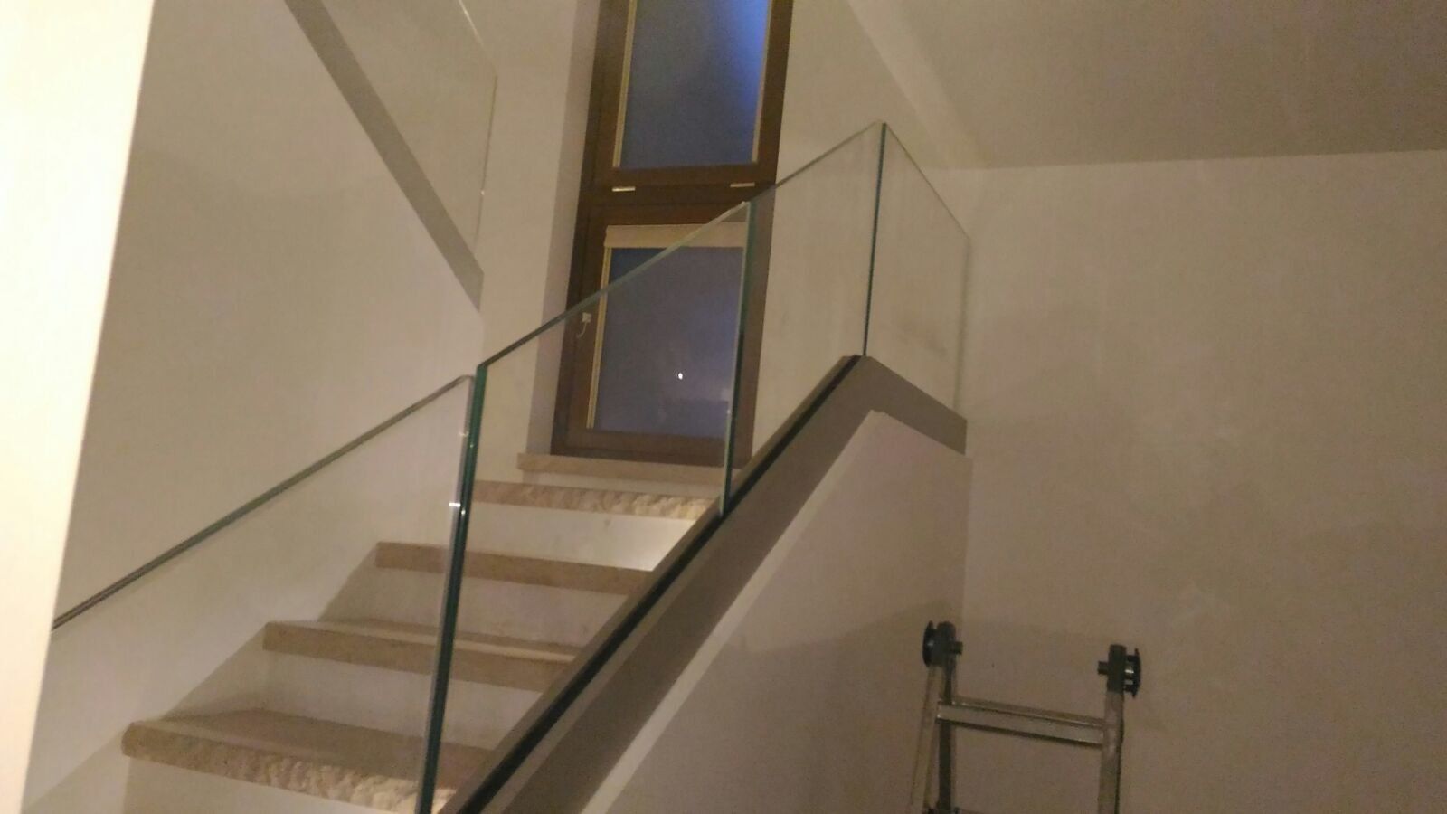 Scala con ringhiera in vetro e porta in cima. Gradini beige, pareti bianche e una scala a pioli.