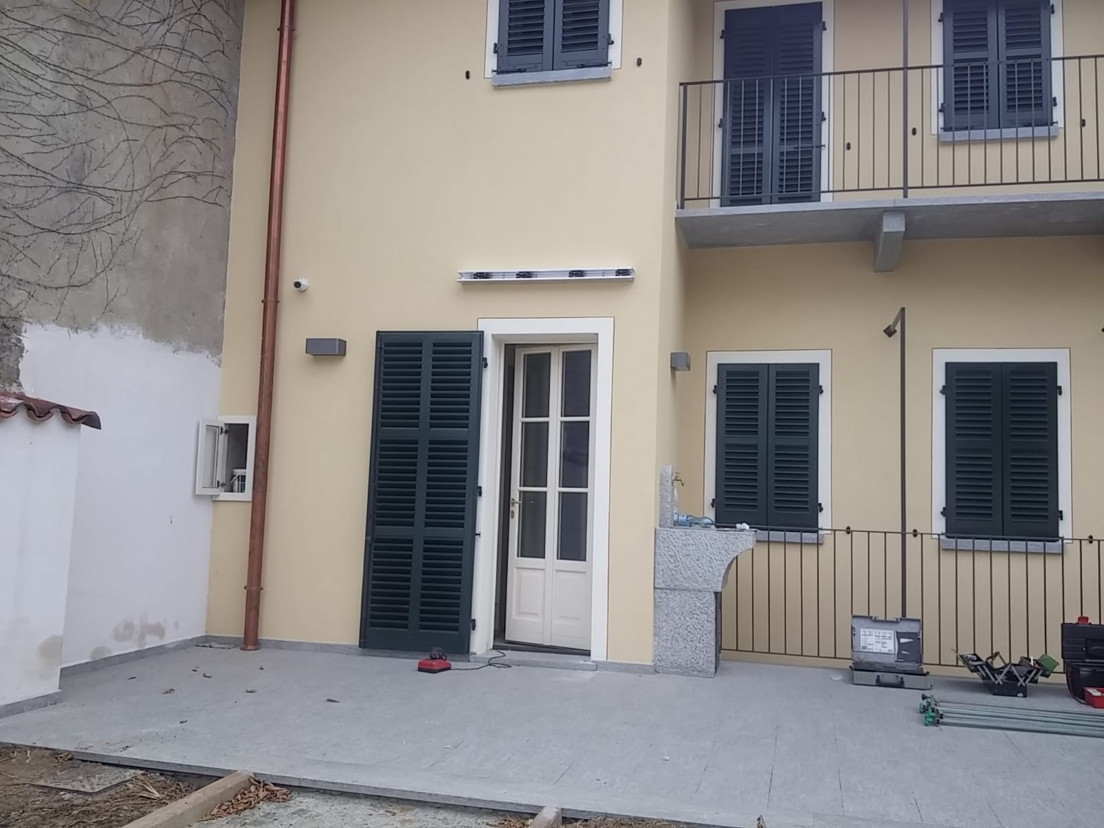 Edificio beige con persiane verdi, balcone e porta aperta su un patio in cemento.