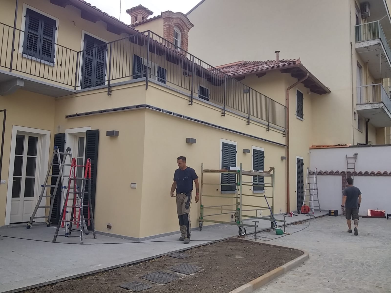 Operai in un cantiere edile; un edificio giallo con impalcature e scale. Due uomini attraversano un cortile.