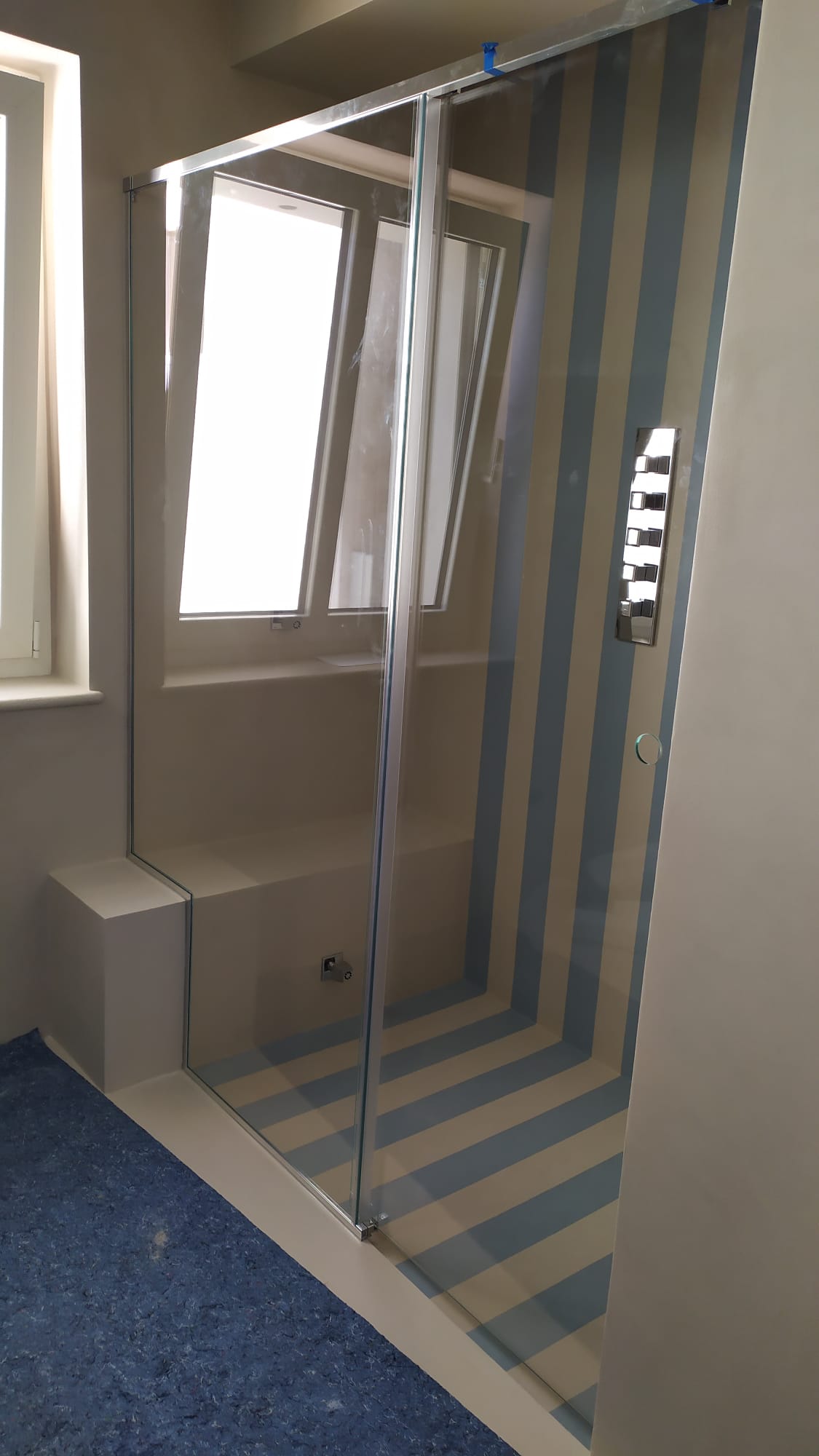 Doccia con porta in vetro, parete a strisce blu e bianche, piccola panca, finestra.