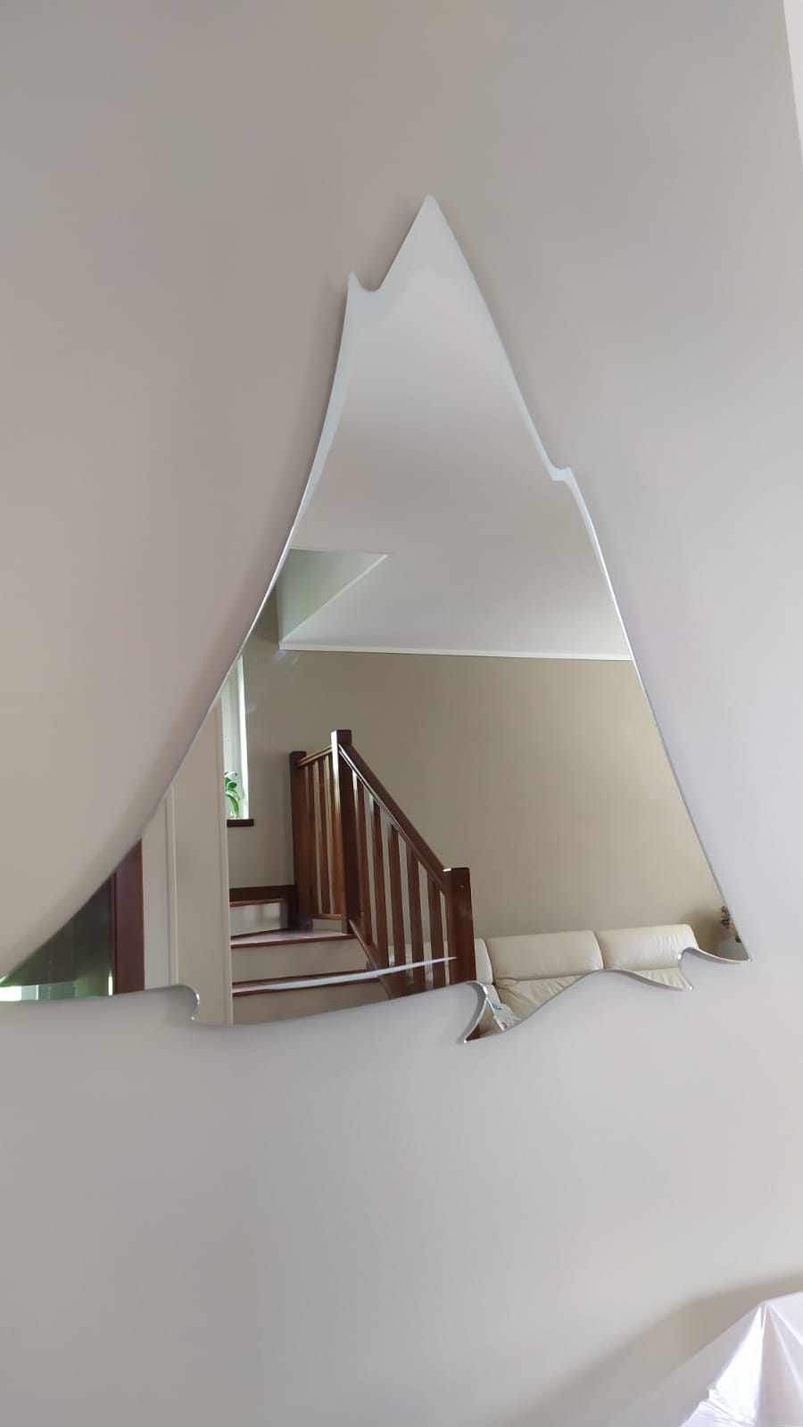 Specchio a forma di montagna appeso a una parete beige; riflette una scala, un divano bianco e un'area verde.
