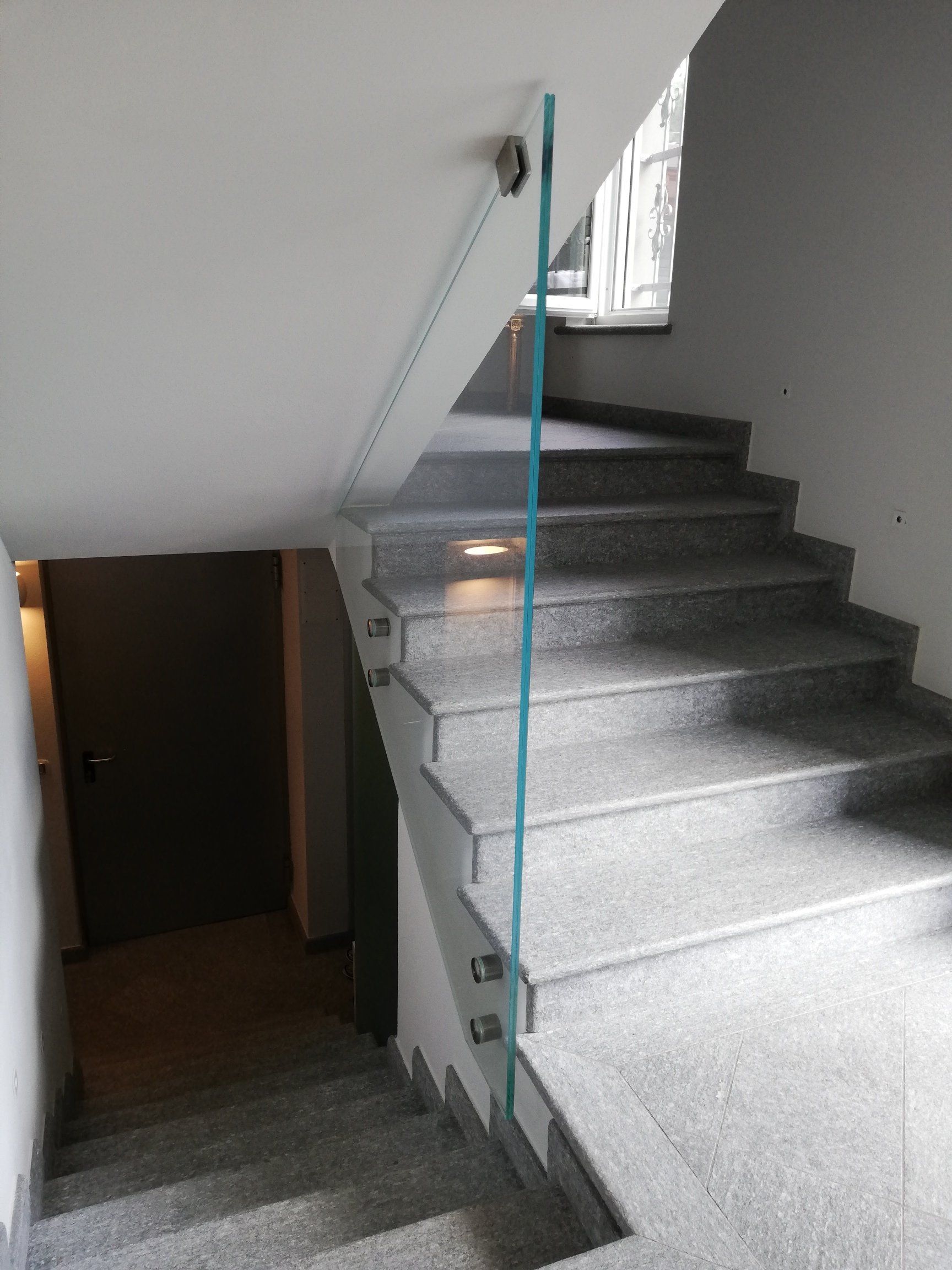 Tromba delle scale con gradini in granito grigio, ringhiera in vetro e una porta scura che conduce a un corridoio.