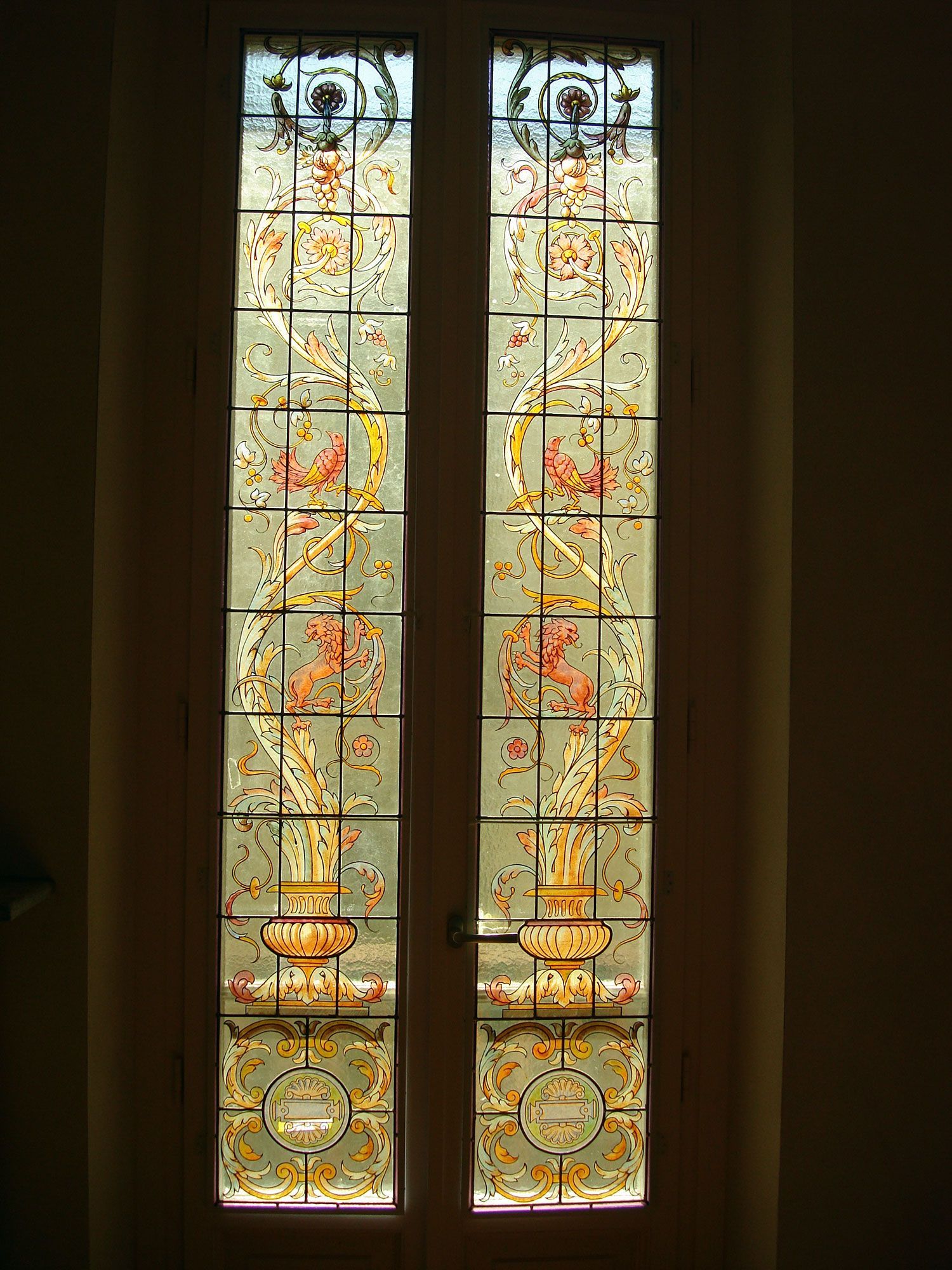 Vetrata alta e decorata con motivi floreali e geometrici in arancione, oro e verde su sfondo trasparente.
