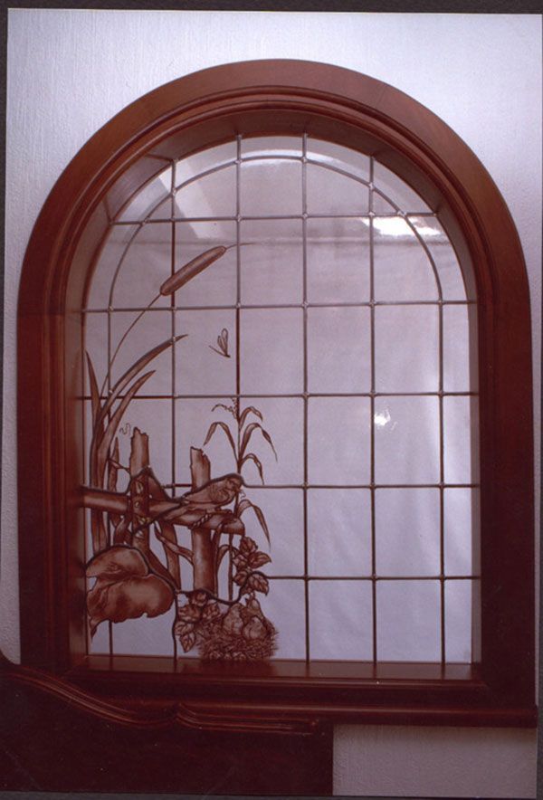 Vetrata con cornice in legno ad arco raffigurante un uccello su una staccionata tra canne e fiori.