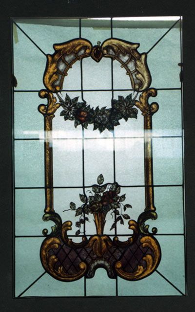 Pannello in vetro colorato con dettagli dorati, bordeaux e floreali; una corona floreale centrale e un vaso.