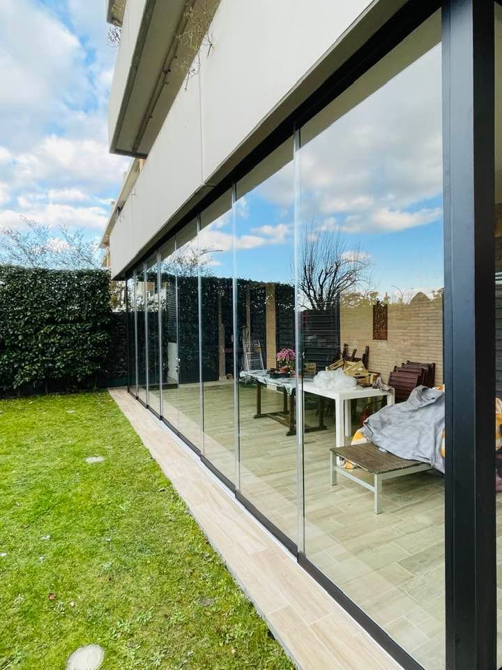 Portefinestre con pareti in vetro in una casa moderna, che riflettono il cielo azzurro e una zona pranzo all'aperto.