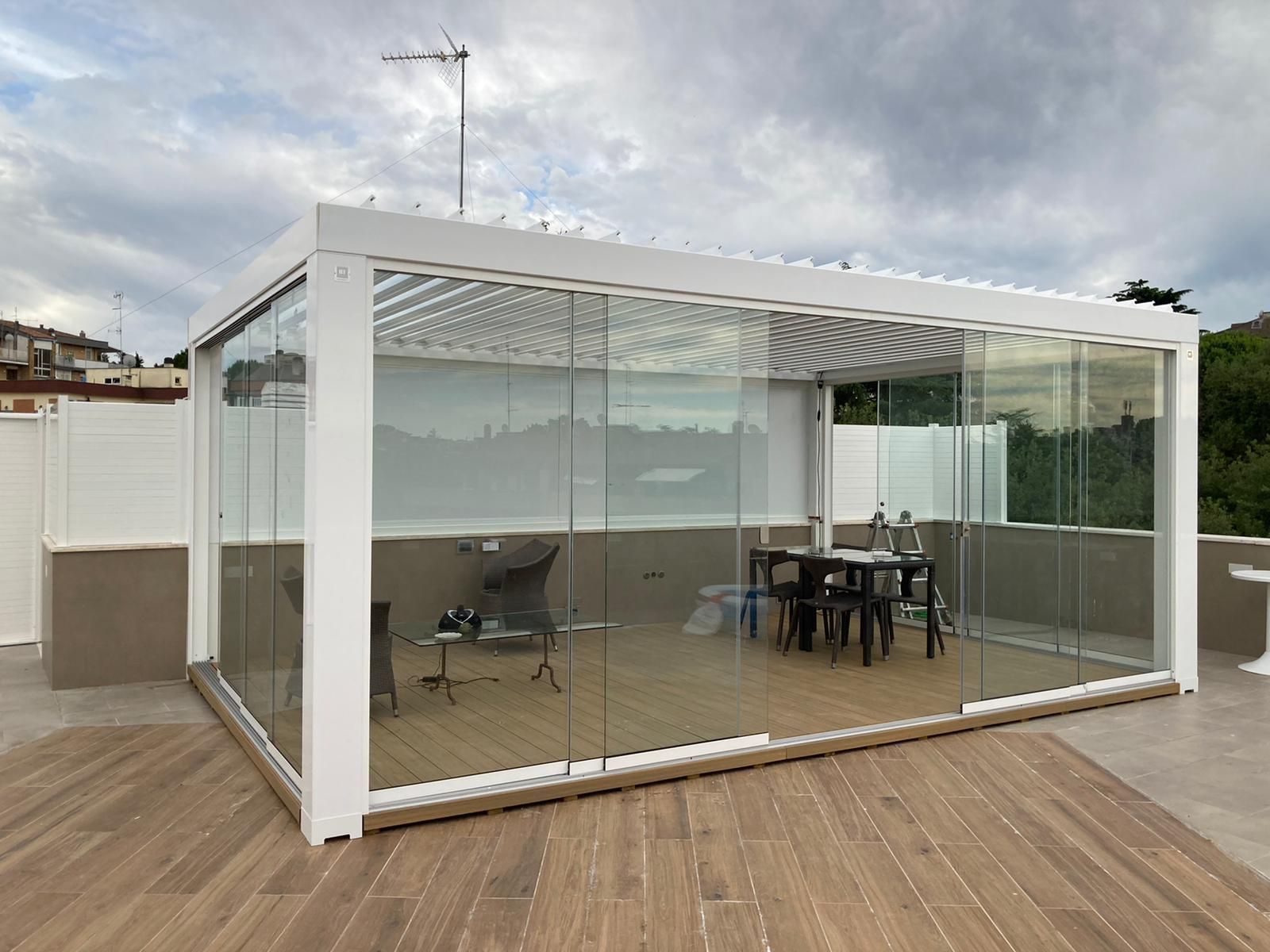 Pergola con pareti in vetro e cornice bianca su una terrazza in legno, con tavolo da pranzo e sedie all'interno.