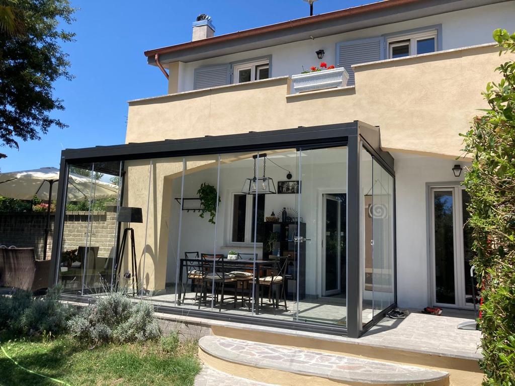 Una moderna veranda con pareti in vetro e un set da pranzo adiacente a una casa beige a due piani.