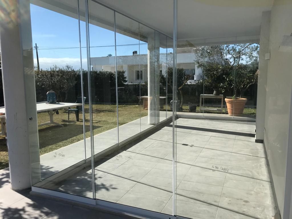 Patio con vetrate che si affaccia su un cortile con alberi, un tavolo e una casa in una giornata di sole.