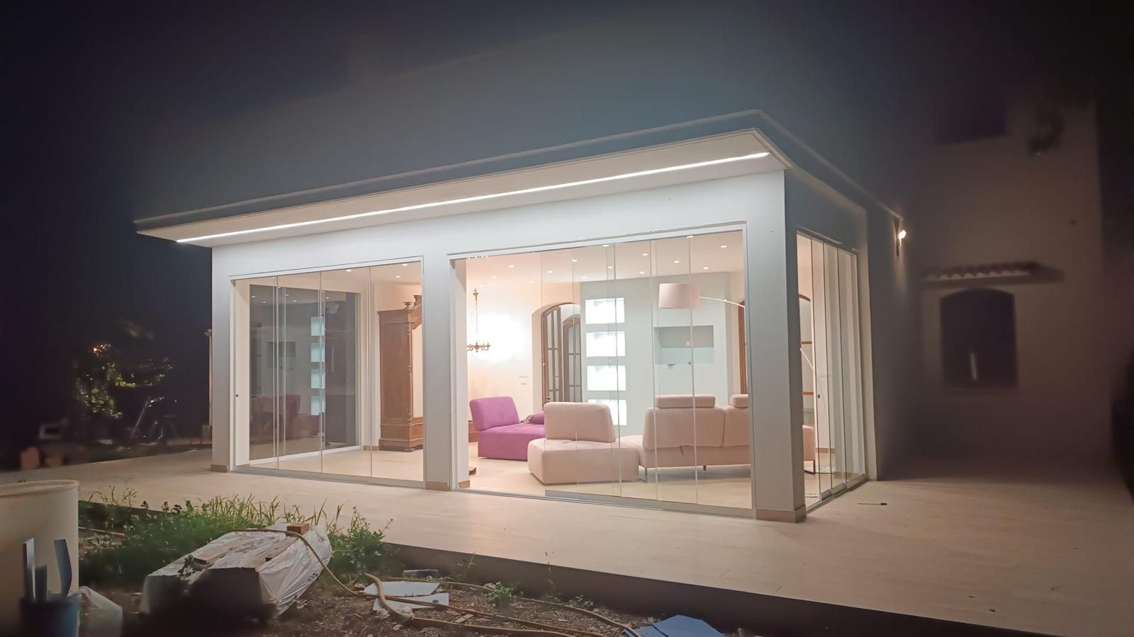 Patio con pareti in vetro di notte, l'interno illuminato rivela mobili rosa e beige. Edificio bianco, cielo scuro.
