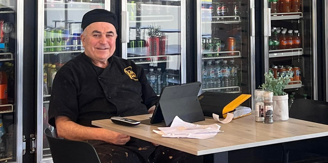 King of The Roast Founder Tony sits at a table with papers, a tablet, and plants, in front of glass-door coolers.