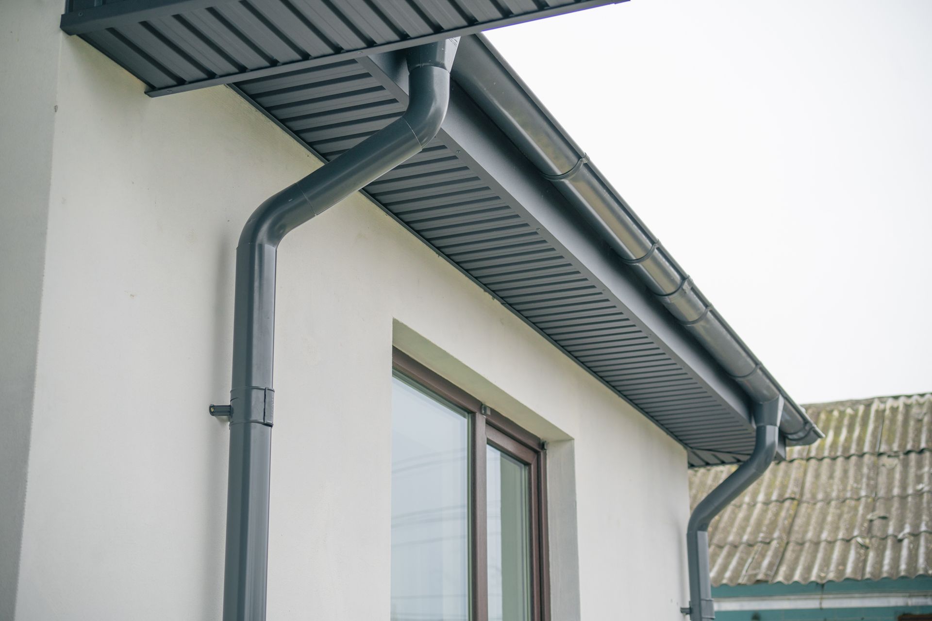 Dark gray rain gutter system on a white building exterior, under a gray overhang.