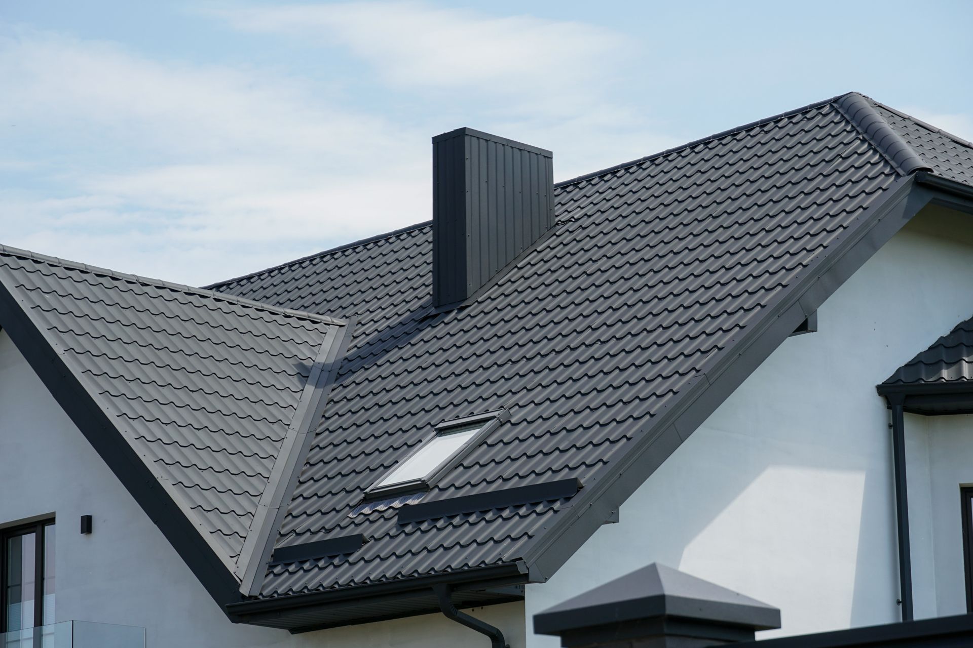 Dark gray metal roof on a white house with a black chimney and a skylight against a blue sky.