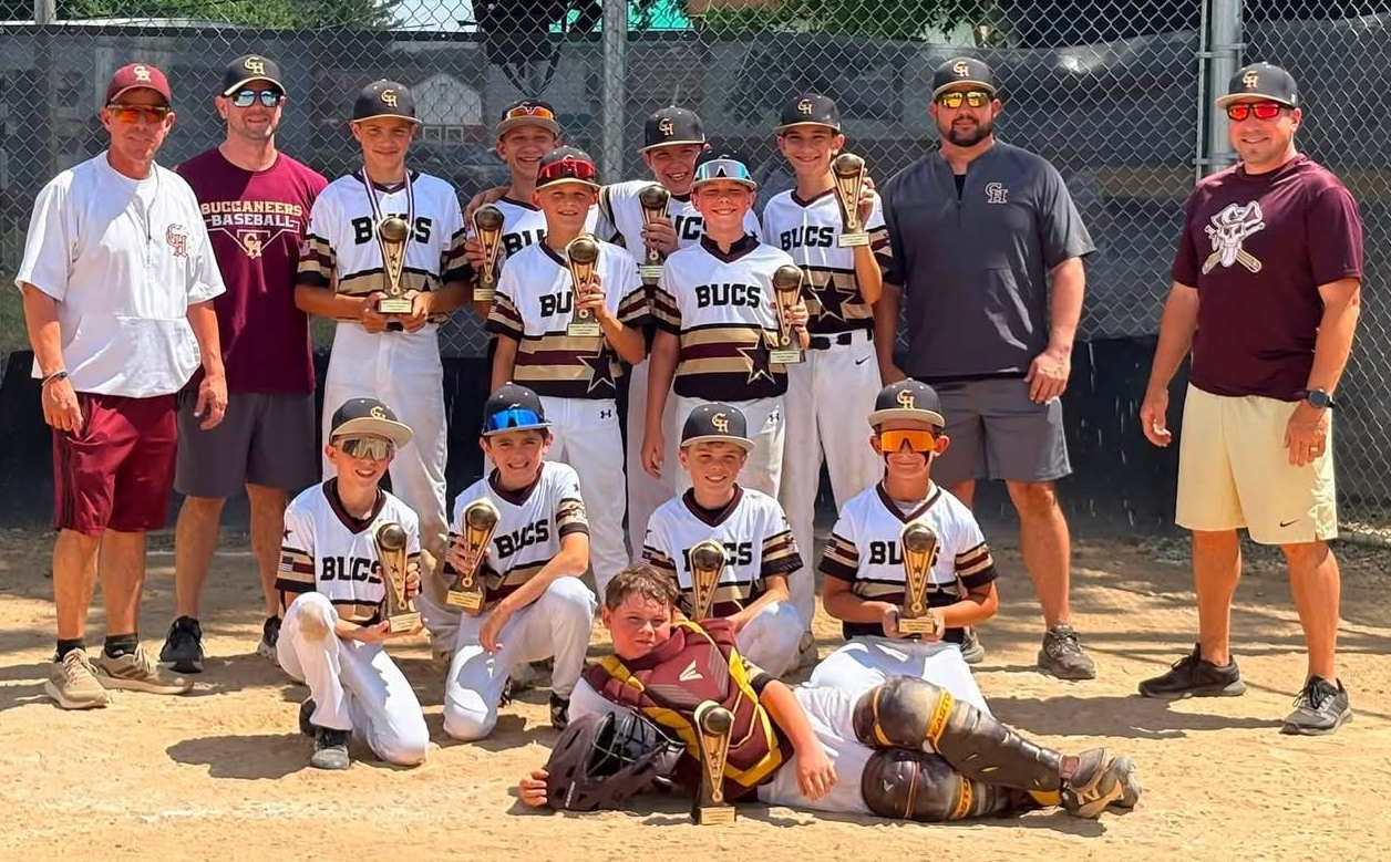 Youth baseball team posing with coaches on a field, wearing white and black uniforms with championship trophy in front