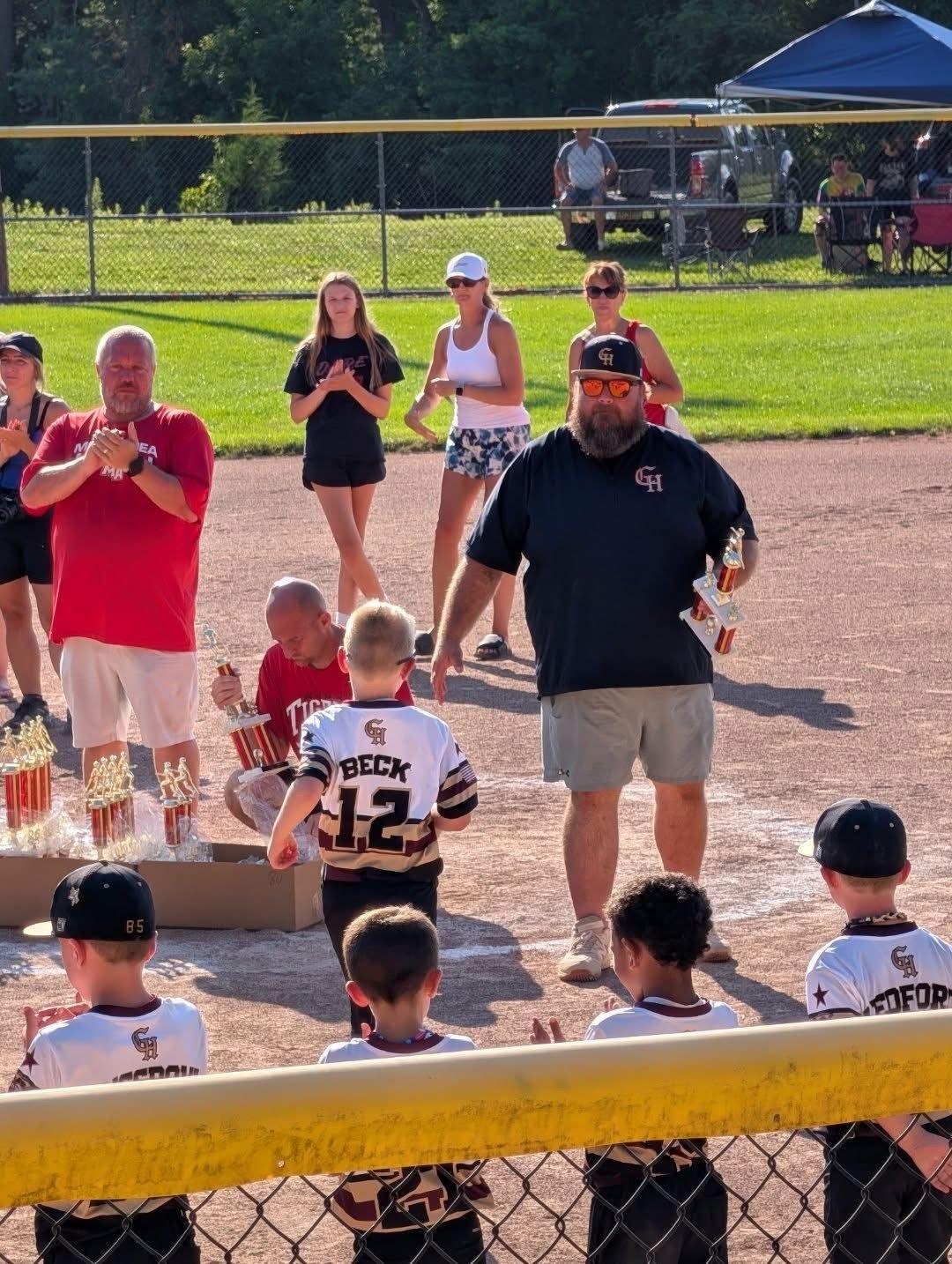 Youth baseball player walks toward home plate while spectators applaud near the field.