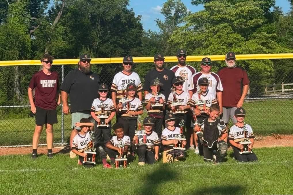 Youth baseball team and coaches posing on a field in front of a yellow fence, with trophy cups displayed in front.