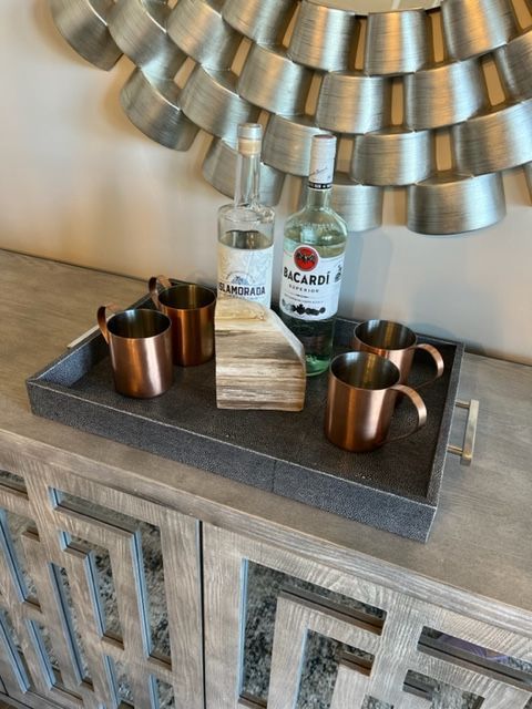 Copper mugs, liquor bottles, and a decorative piece on a tray atop a gray cabinet, with a silver mirror in the background.