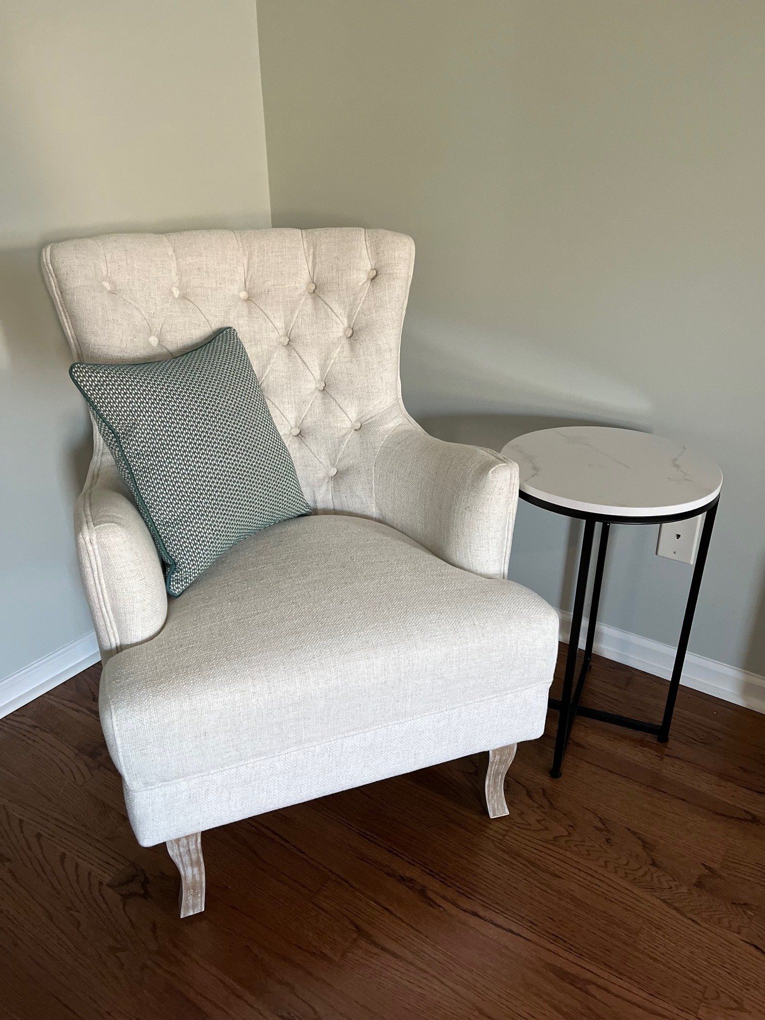 Cream tufted armchair with a teal pillow, next to a small round table, in a corner of a room.