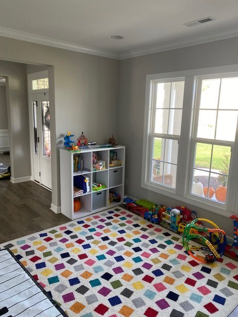 A child's playroom with a colorful rug, white shelving unit, and toys near a window.