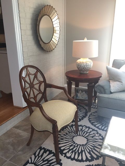 Oval-backed wooden chair with cream cushion, round mirror, and lamp on a side table, all on patterned rug.