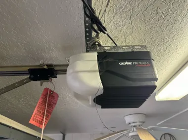 Garage door opener unit mounted on a ceiling, with a red tag hanging from the arm.