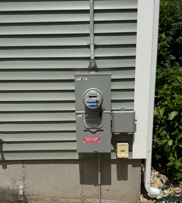Gray electrical meter and service box mounted on house exterior with conduit and louvered siding.