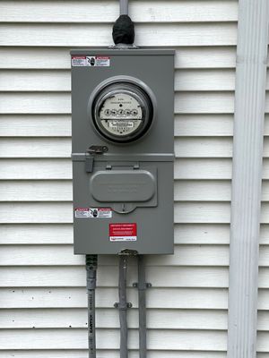 Gray electric meter box mounted on white siding with conduit pipes below