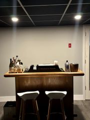 Small bar cart with bottles and two stools against a plain wall under ceiling lights