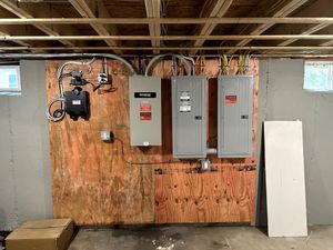 Basement wall with electrical panels, wiring, and a small window; plywood board leaning on the right.
