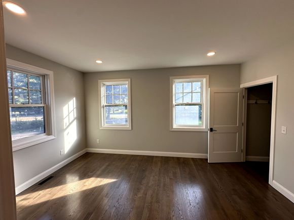 Empty beige room with hardwood floors, three windows, and an open door letting in sunlight.