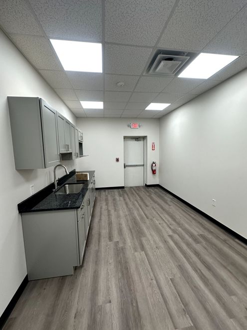 Empty break room with gray flooring, white walls, cabinets, countertop, and ceiling lights.