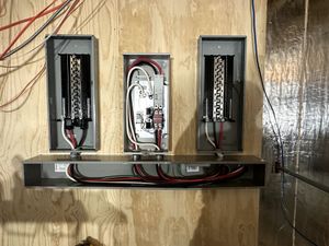 Three open electrical breaker panels mounted on a plywood wall with exposed wiring.