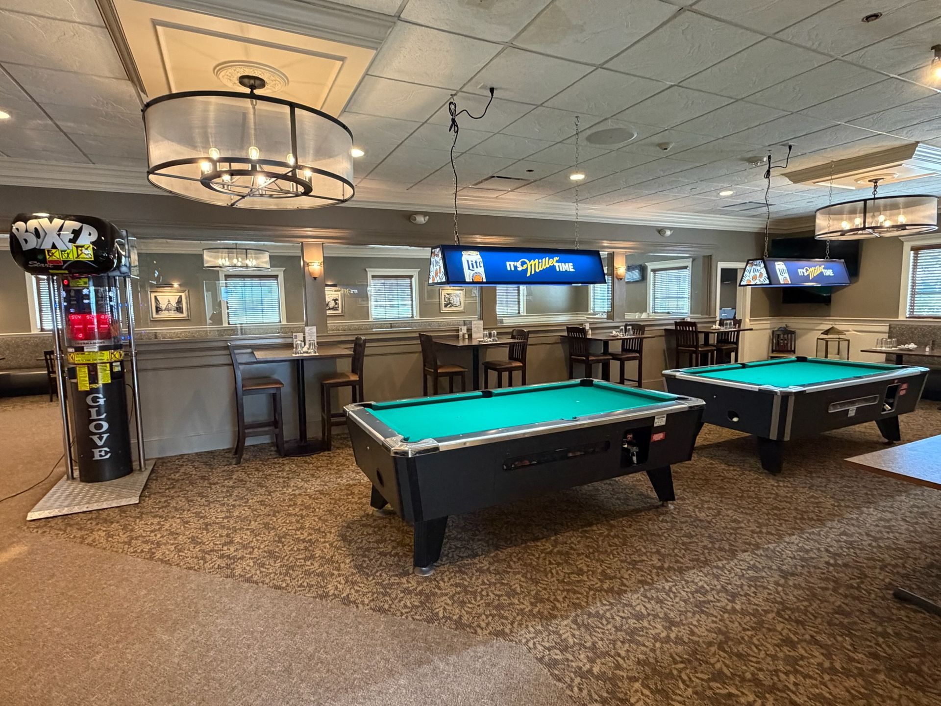 Pool hall with two green felt tables, bar seating, TVs, and a carpeted ceiling-lit interior