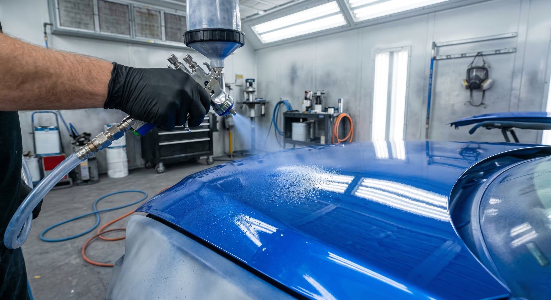 A person in black gloves uses a spray gun to apply blue paint to a car hood inside a professional auto body shop.