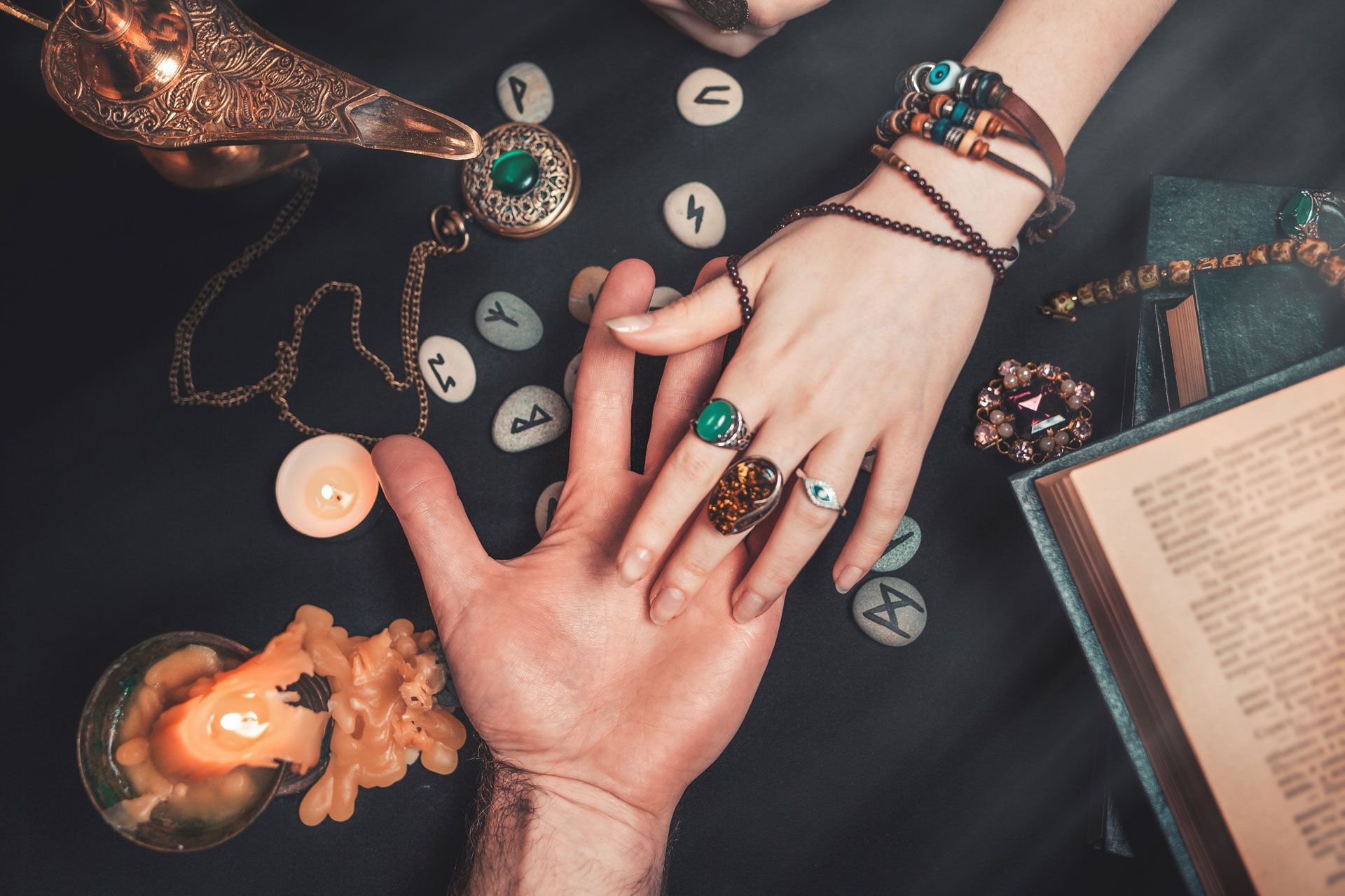 Hands of two people, palm-to-palm, surrounded by runes, candles, and jewelry on a dark table.