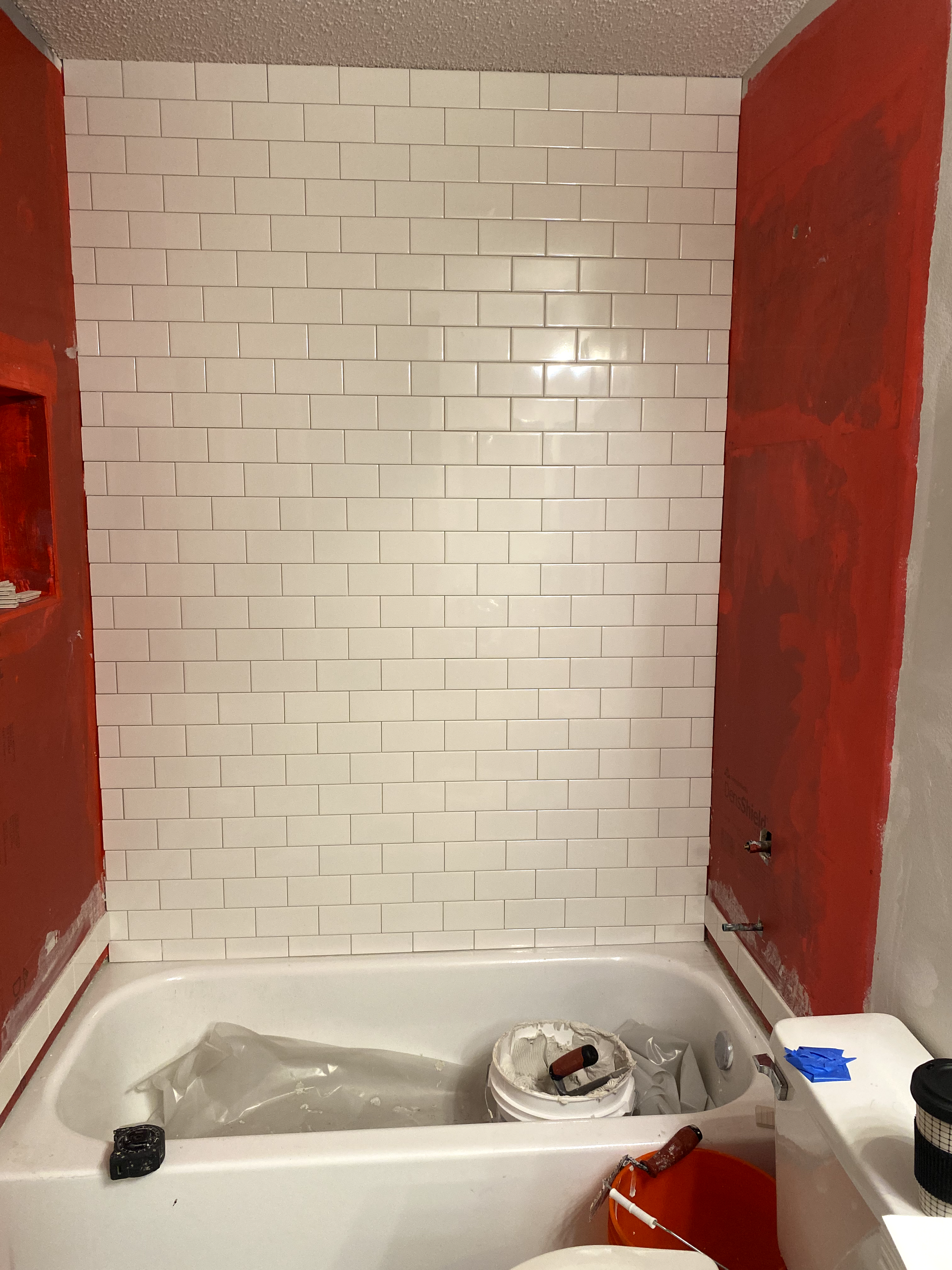 White tiled shower wall above a bathtub; red walls on sides. Construction materials visible.