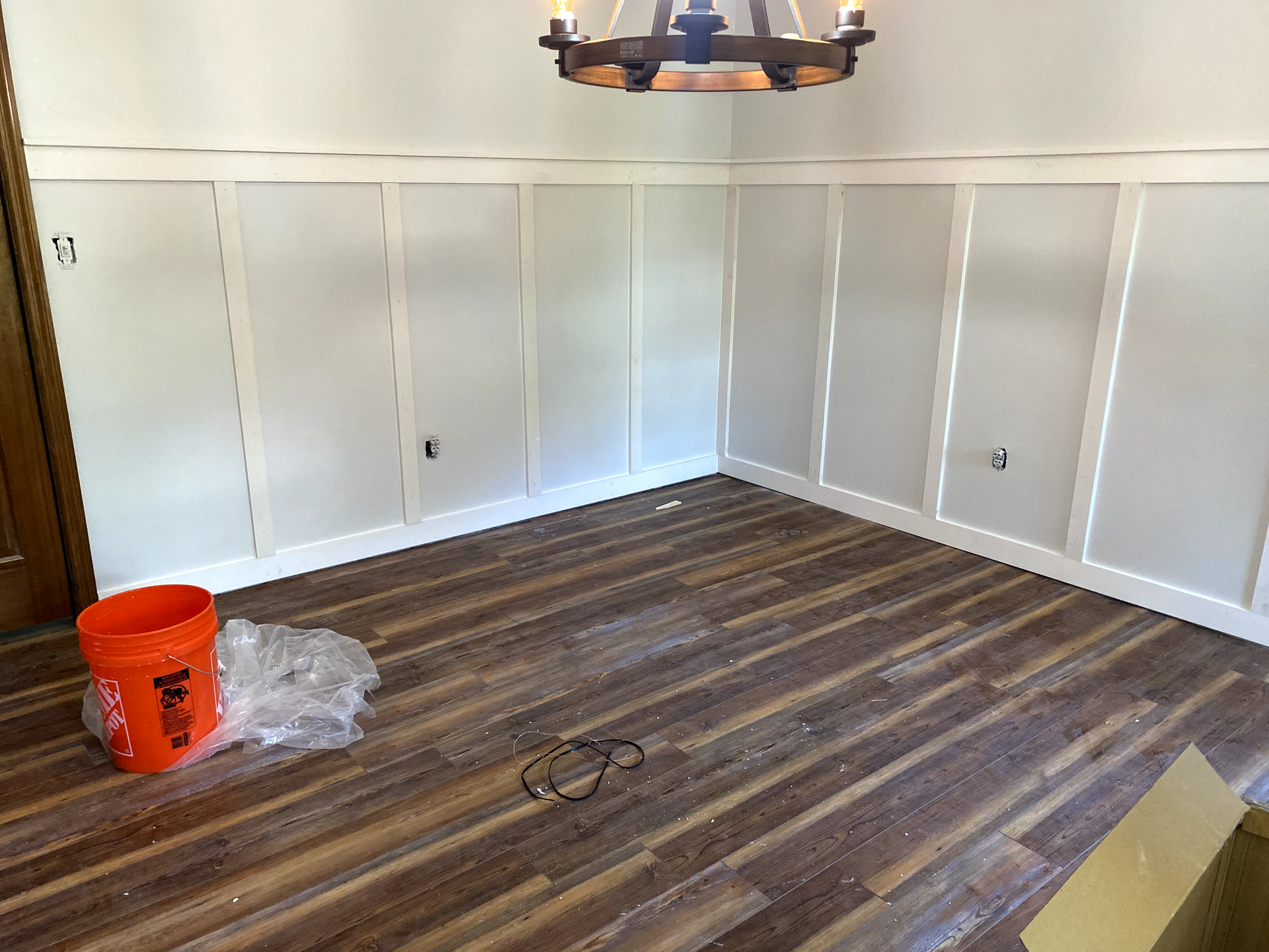 Empty room with wood-look flooring, light gray and white paneled walls, a chandelier, and a construction bucket.