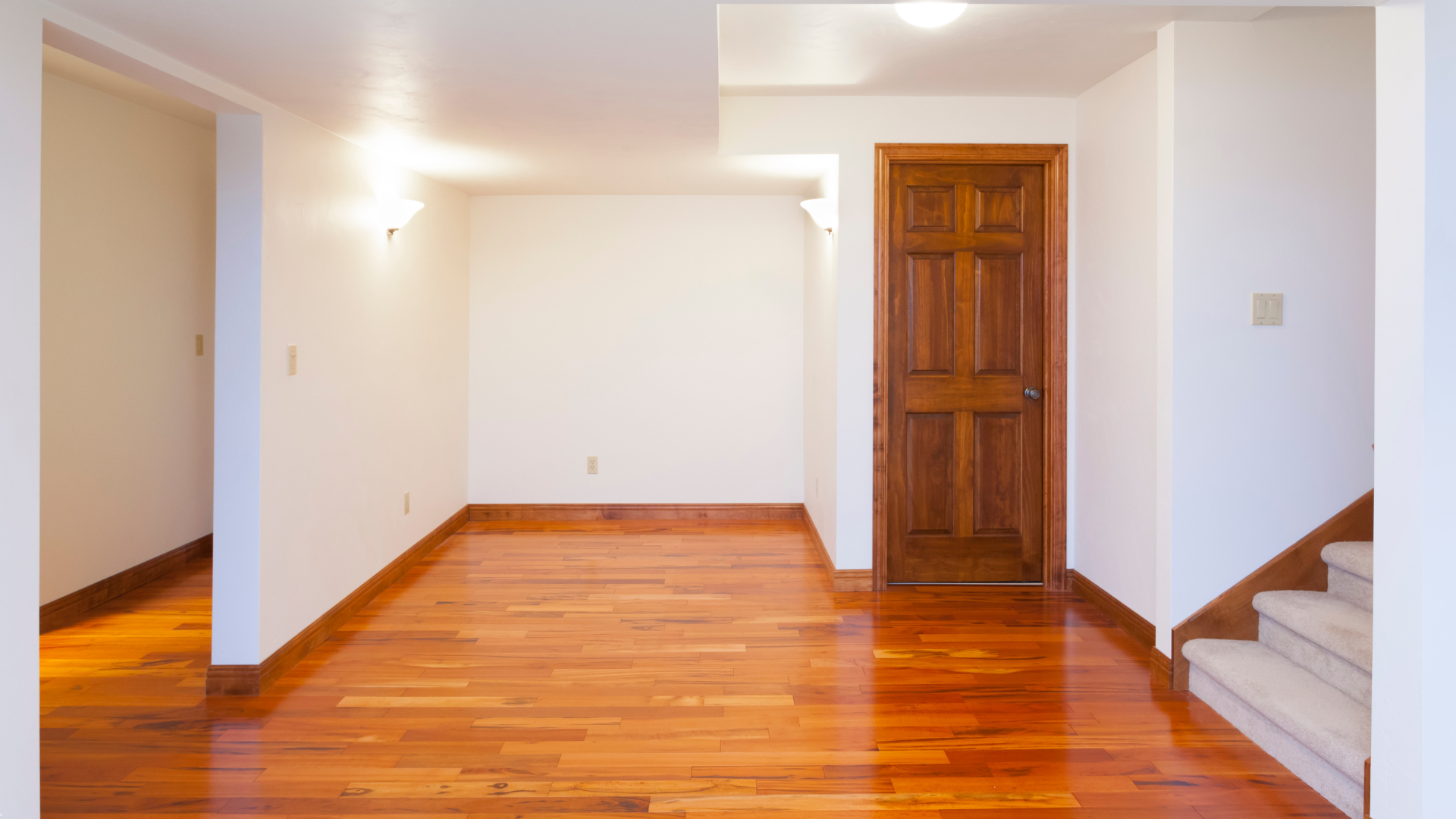 Empty room with hardwood floors, white walls, wooden door, and a staircase.