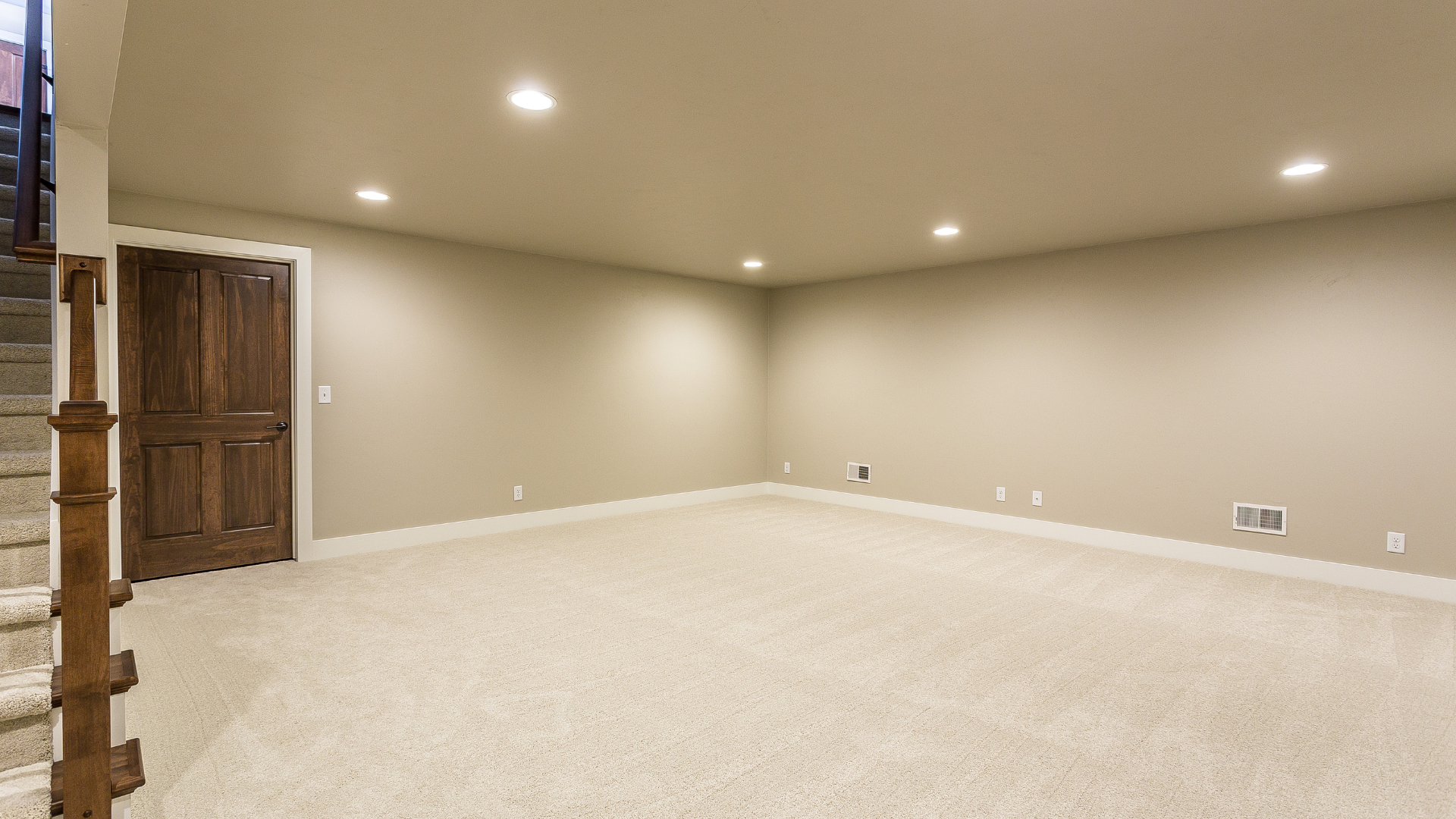 Empty, carpeted basement room with a wooden door, beige walls, and recessed lighting.