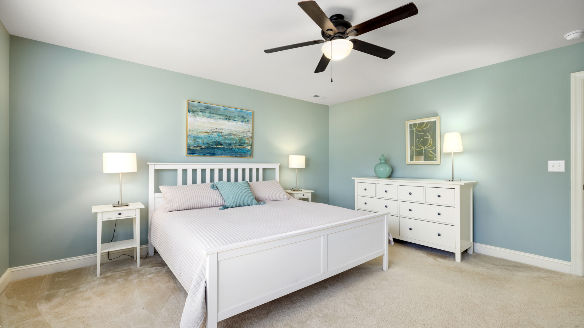 Bedroom with a white bed, dresser, and nightstand. Light blue walls and beige carpet. Ceiling fan.