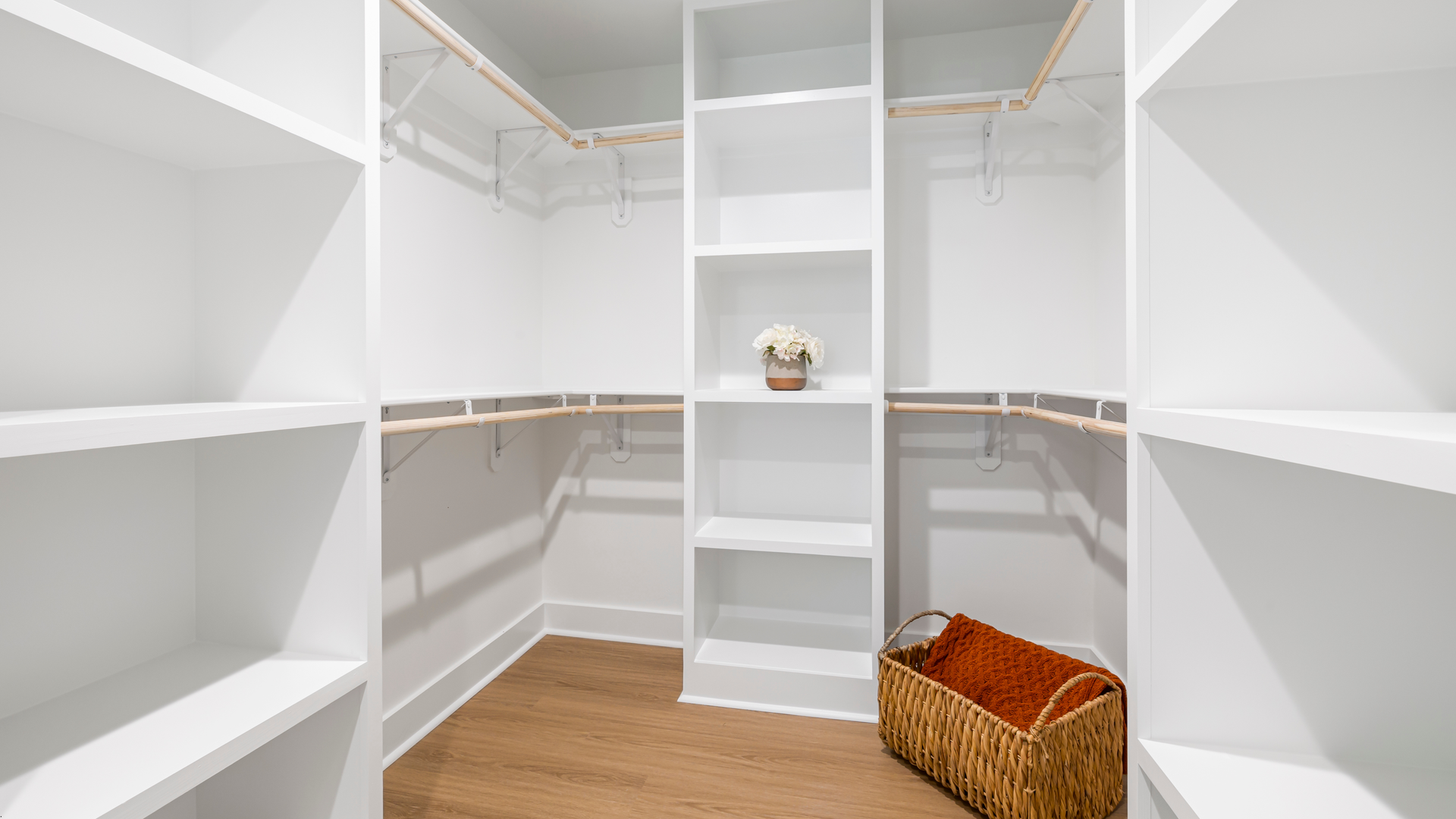 Empty white walk-in closet with shelves, hanging rods, and a woven basket on a wood floor.