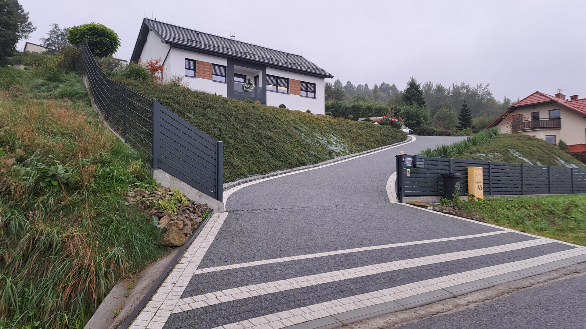 A paved driveway leads to a house on a hillside. The house is white with a dark roof.