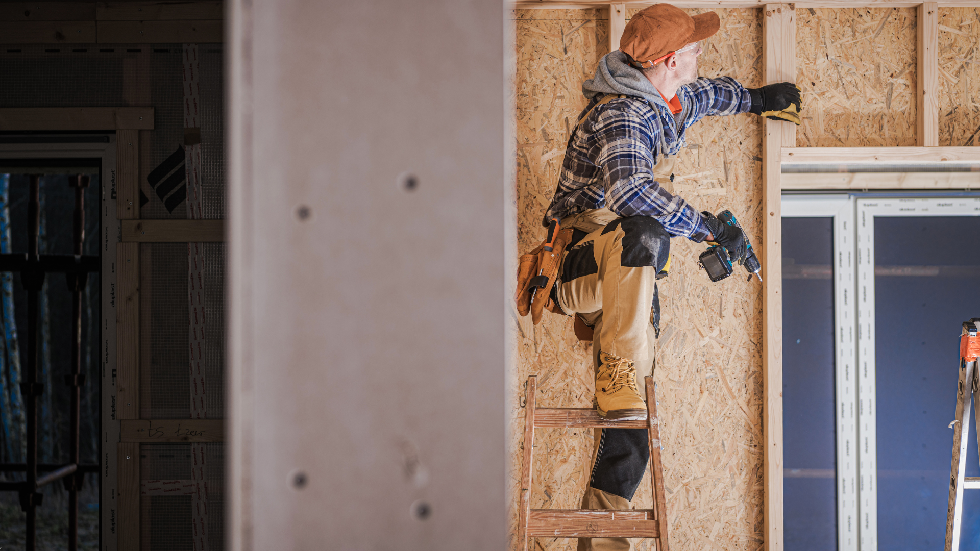 Carpenter on a ladder, using a power drill to frame a window in a wood-paneled construction site.