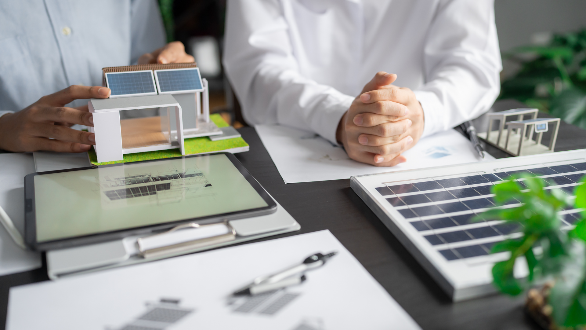 Two people reviewing house model with solar panels. Documents, tablet, and pen on the table.