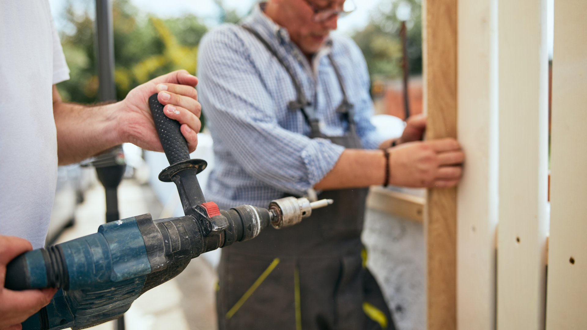 Person holding drill, assisting another person constructing wooden structure outdoors.