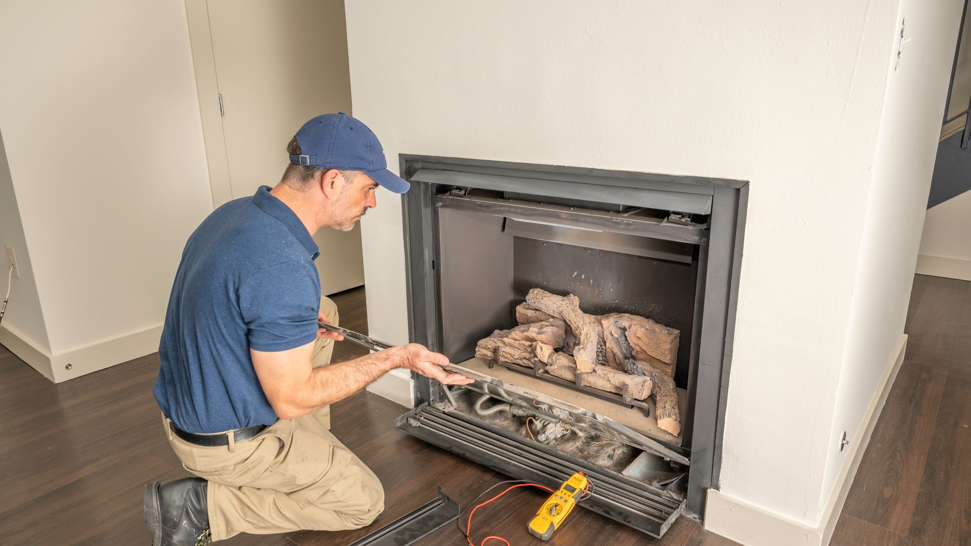 Man in blue shirt and cap kneeling in front of a fireplace, inspecting the gas logs.