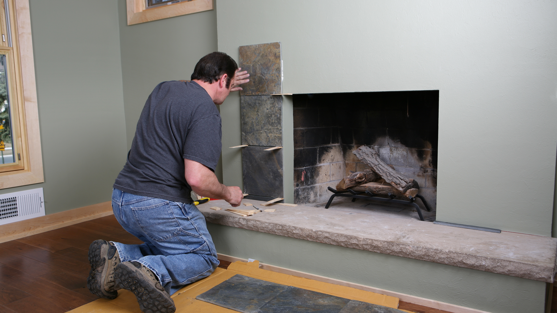 Man kneeling, tiling fireplace wall. He is wearing a blue shirt and jeans. Green wall. Wood floor.
