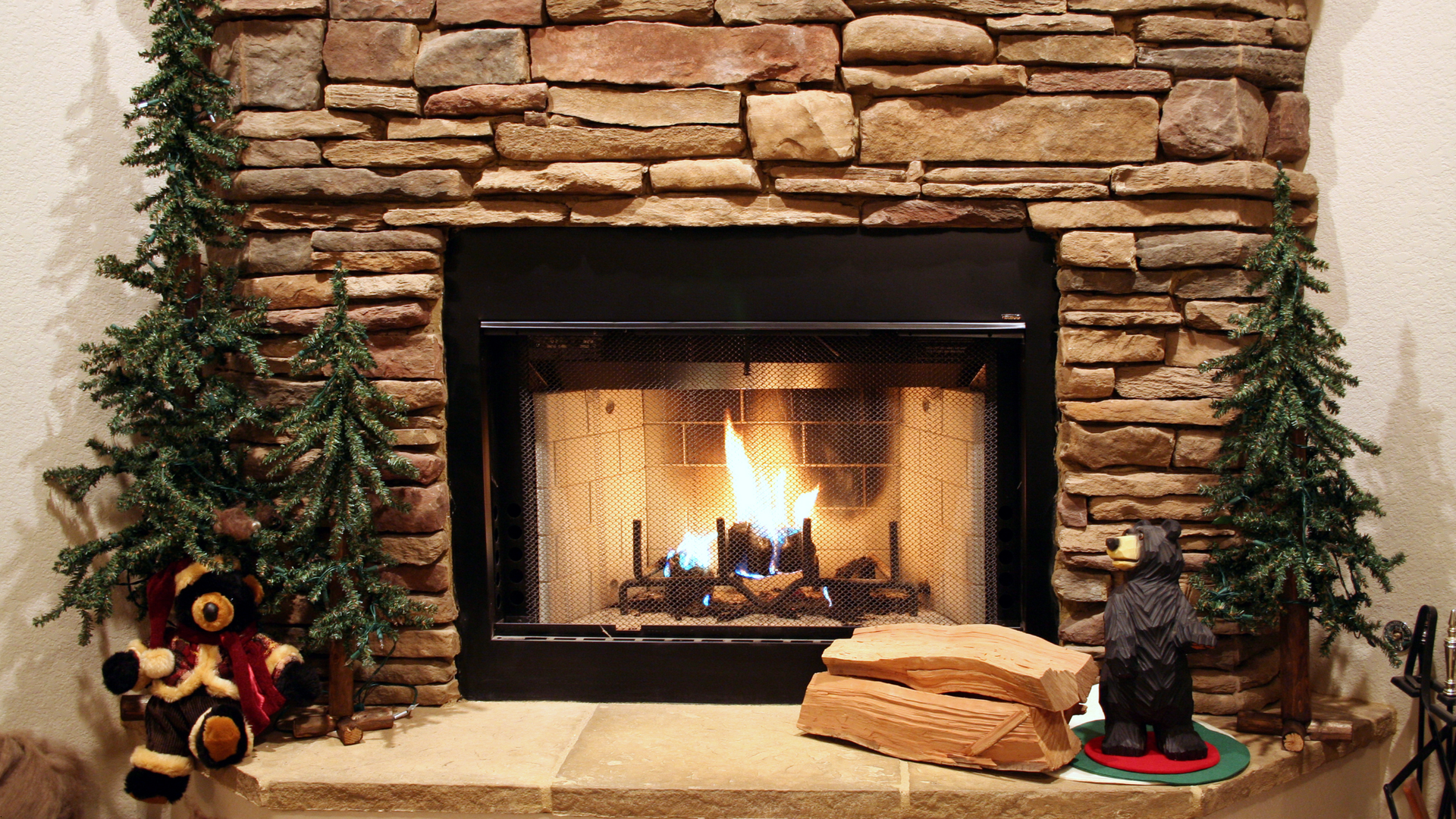 Fireplace with burning fire, flanked by two small Christmas trees and a bear statue.